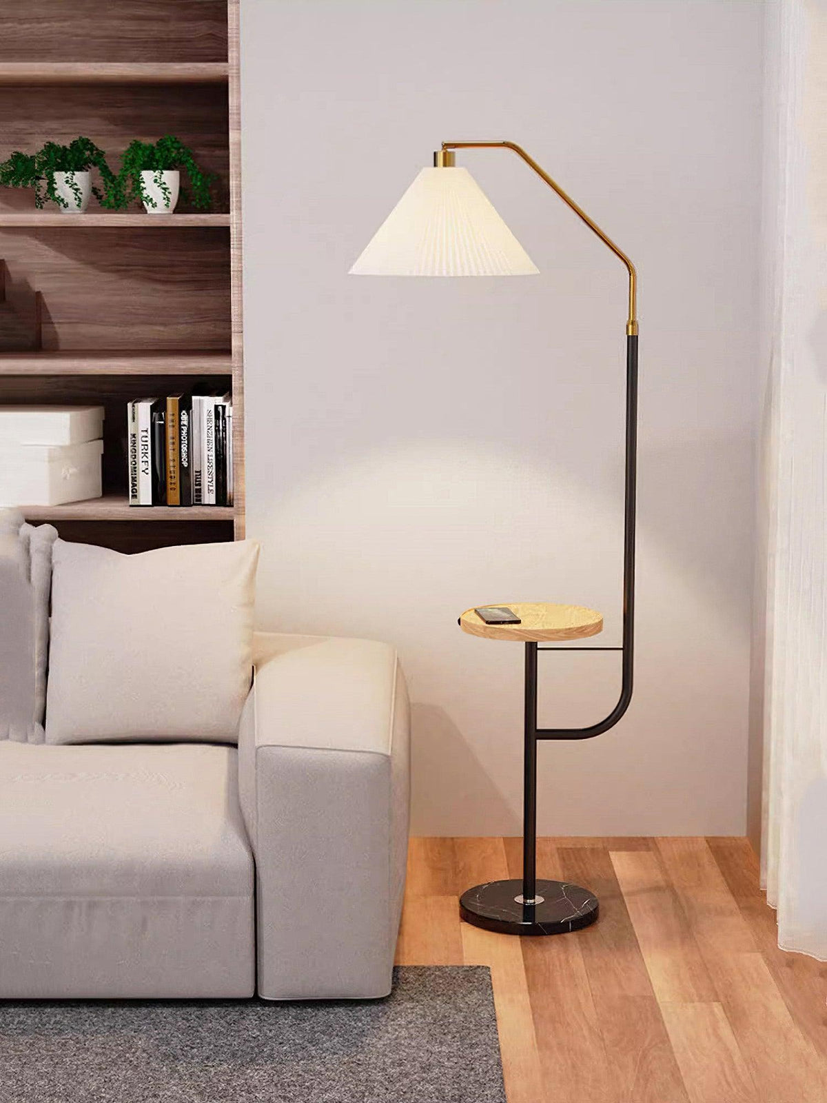 Clérachic floor lamp against the same bookshelf, cream pleated shade lit above a wood tray holding a phone, grey rug underfoot.