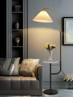 Clérachic floor lamp beside a dark cabinet and a grey sofa with patterned cushions, cream shade lit above a marble tray topped with white flowers.