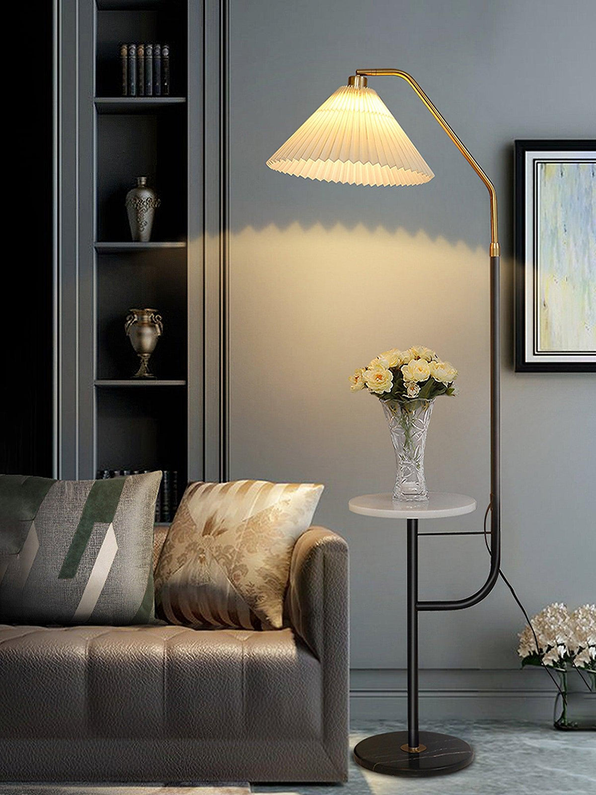 Clérachic floor lamp beside a dark cabinet and a grey sofa with patterned cushions, cream shade lit above a marble tray topped with white flowers.