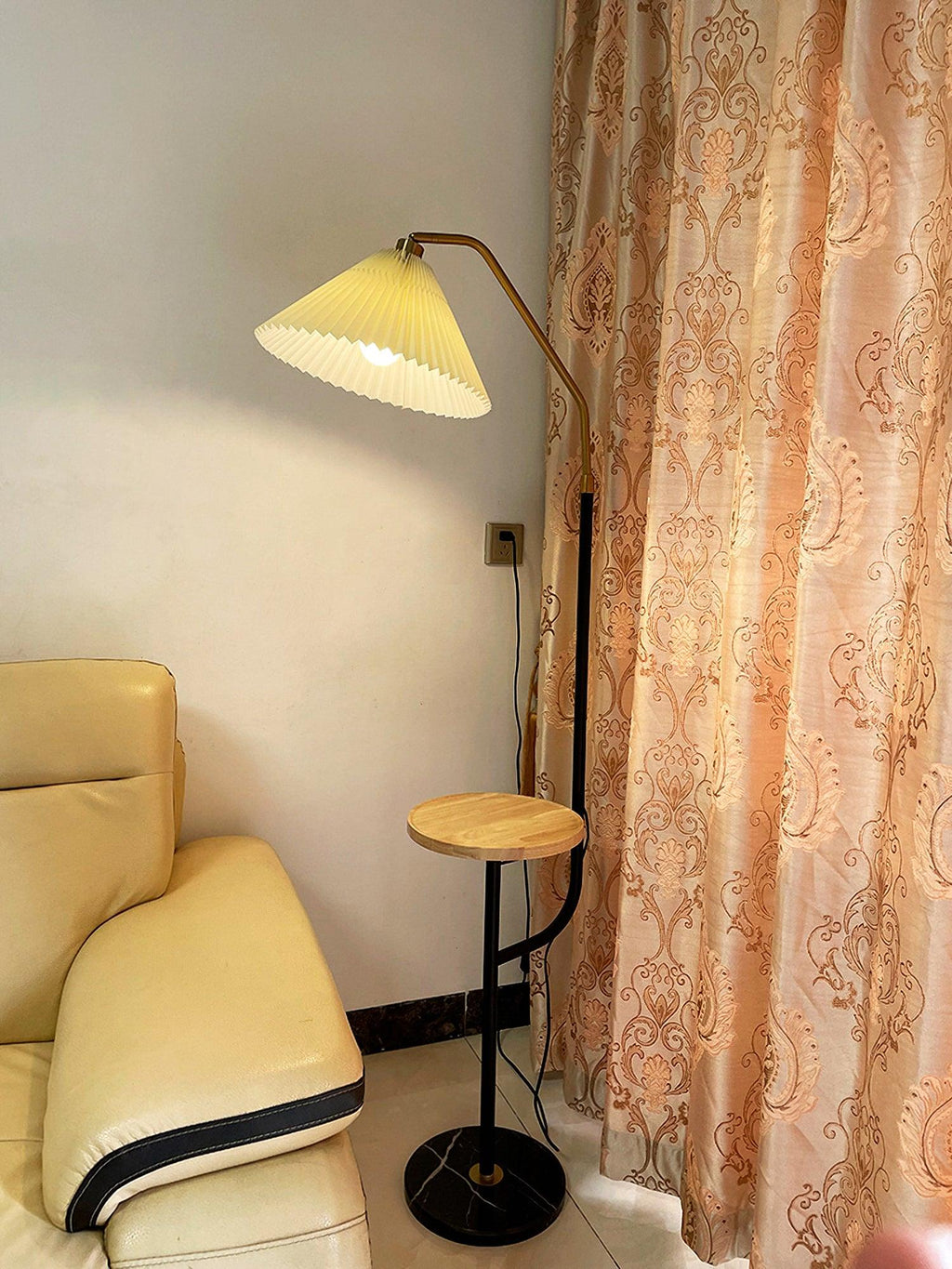 Clérachic floor lamp beside a tan leather recliner and a sheer floral curtain, cream pleated shade lit above a wood tray, cord reaching a wall socket.