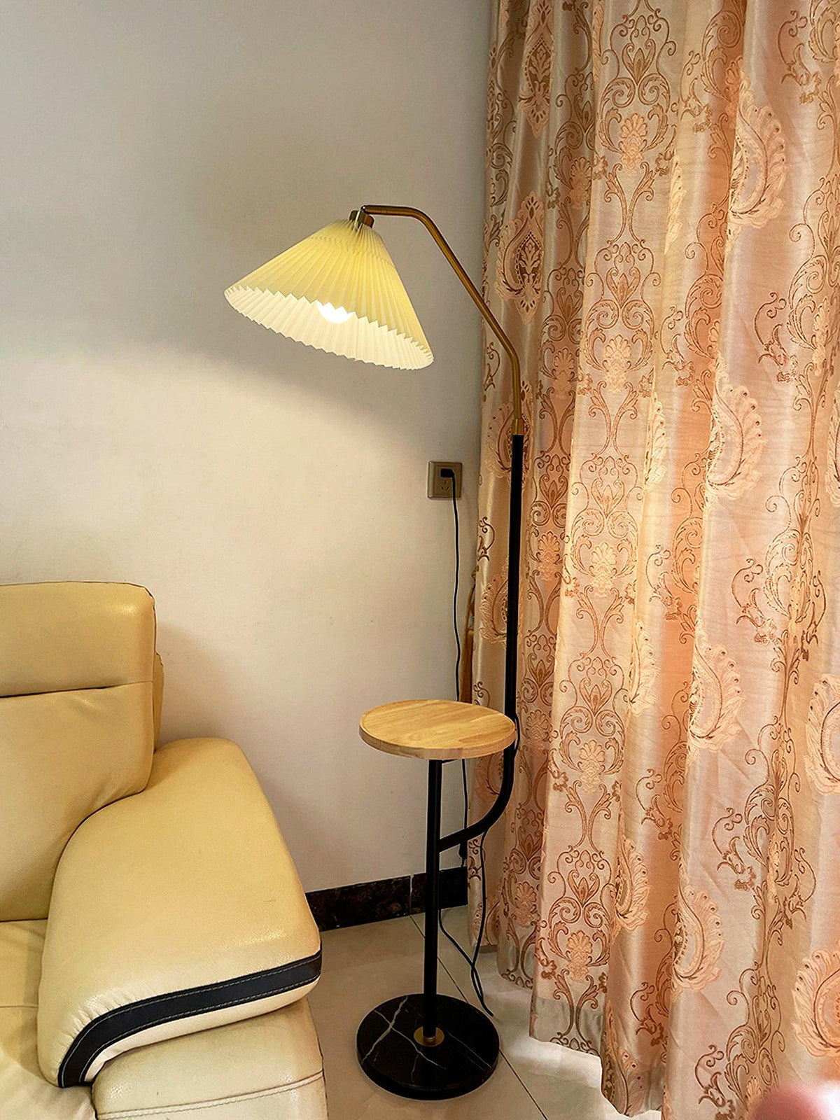 Clérachic floor lamp beside a tan leather recliner and a sheer floral curtain, cream pleated shade lit above a wood tray, cord reaching a wall socket.