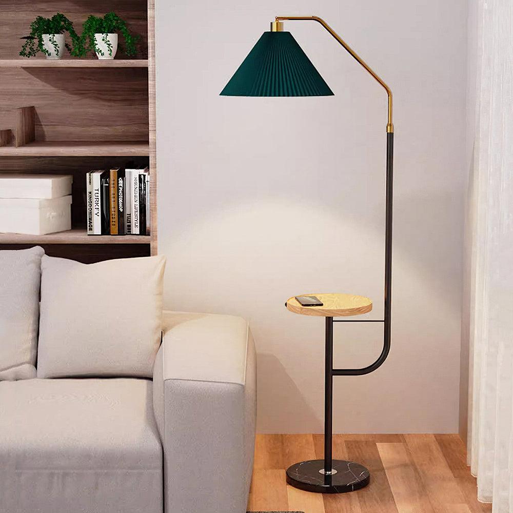 Clérachic floor lamp standing against a bookshelf of stacked books and storage boxes, green pleated shade lit above a wood tray holding a phone.