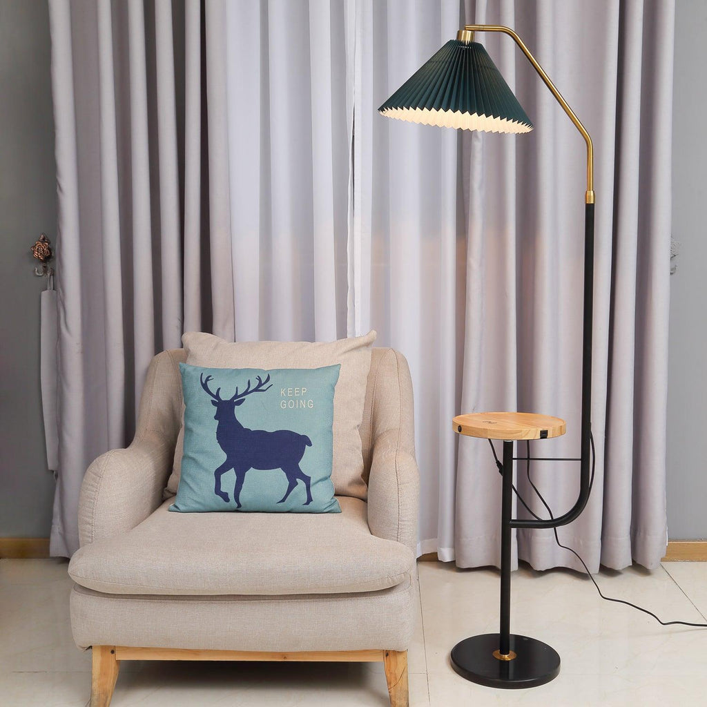 The Clérachic floor lamp lit beside a cream armchair with a blue deer cushion, its green pleated shade glowing above a wood tray holding a teacup.