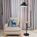 The Clérachic floor lamp lit beside a cream armchair with a blue deer cushion, its green pleated shade glowing above a wood tray holding a teacup.