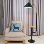The Clérachic floor lamp lit beside a cream armchair with a blue deer cushion, its green pleated shade glowing above a wood tray holding a teacup.