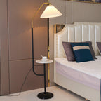 Clérachic floor lamp beside a studded cream leather bed headboard, cream pleated shade lit above a marble tray set with a cup and saucer.