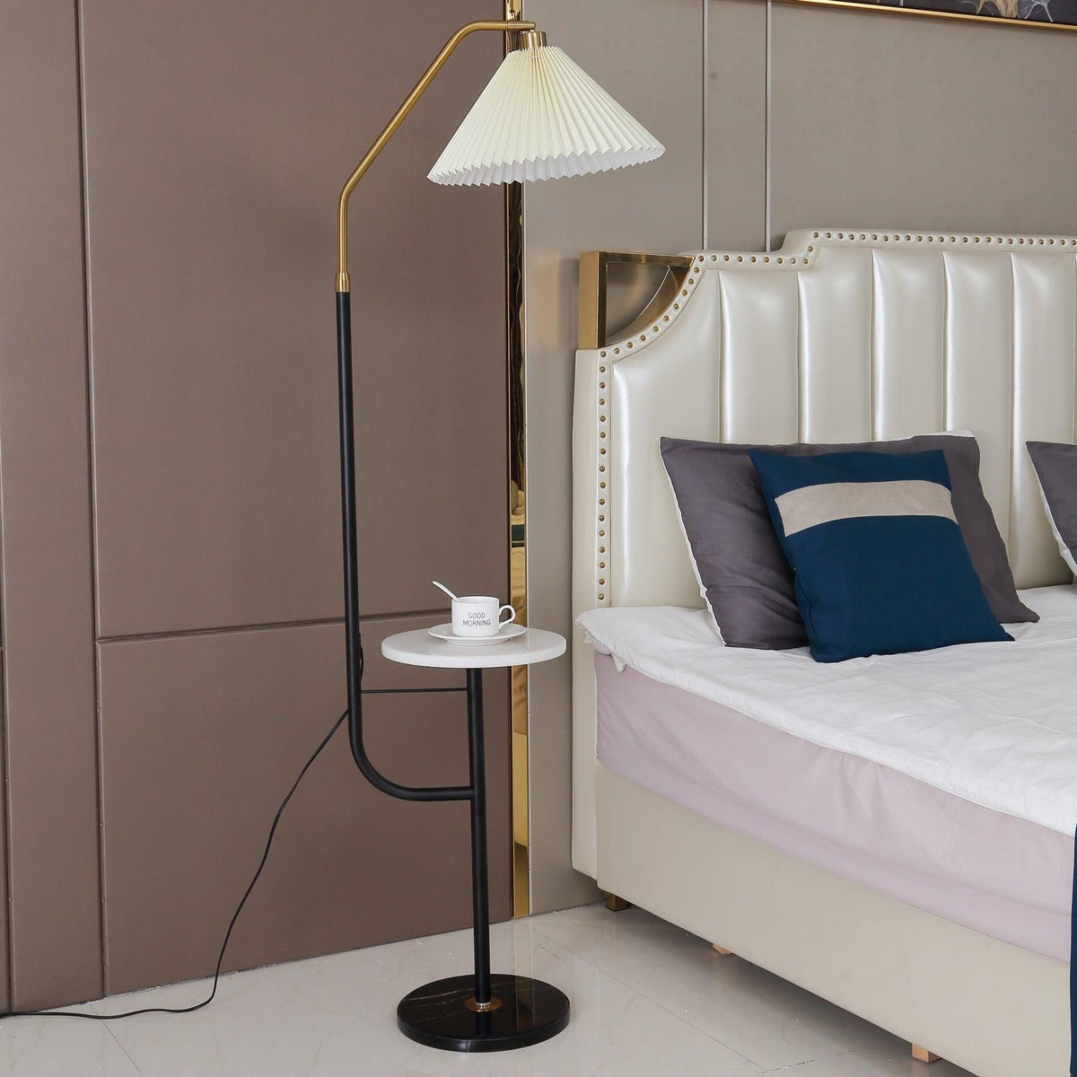 Clérachic floor lamp shown full length beside the same studded headboard, cream shade lit above a marble tray with a cup and saucer.