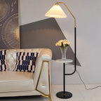 Clérachic floor lamp beside a tufted cream sofa with brass-trimmed legs, green shade lit above a marble tray topped with a small vase of yellow flowers.