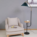 Clérachic floor lamp beside a beige armchair under a framed Paris print, green pleated shade lit above a wood tray set with a cup and saucer.