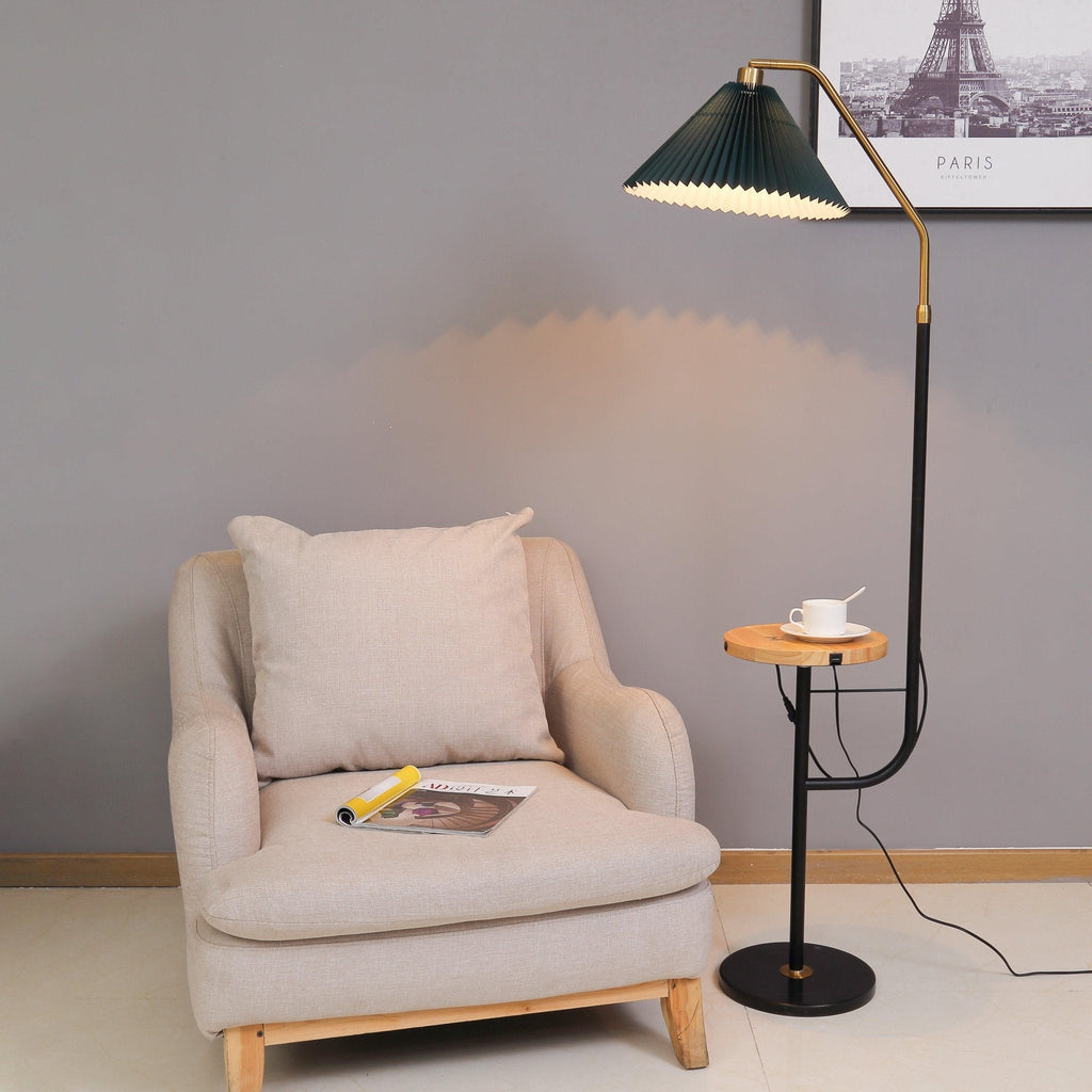 Clérachic floor lamp beside a beige armchair with a magazine on the seat, cream pleated shade lit above a wood tray holding a teacup.