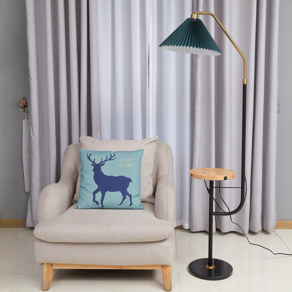 Clérachic floor lamp beside a cream armchair with a blue deer cushion, green pleated shade lit above a wood tray holding a teacup.