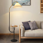 Clérachic floor lamp beside a beige sofa with grey pillows, its cream pleated shade lit warmly above a wood tray holding a teacup.
