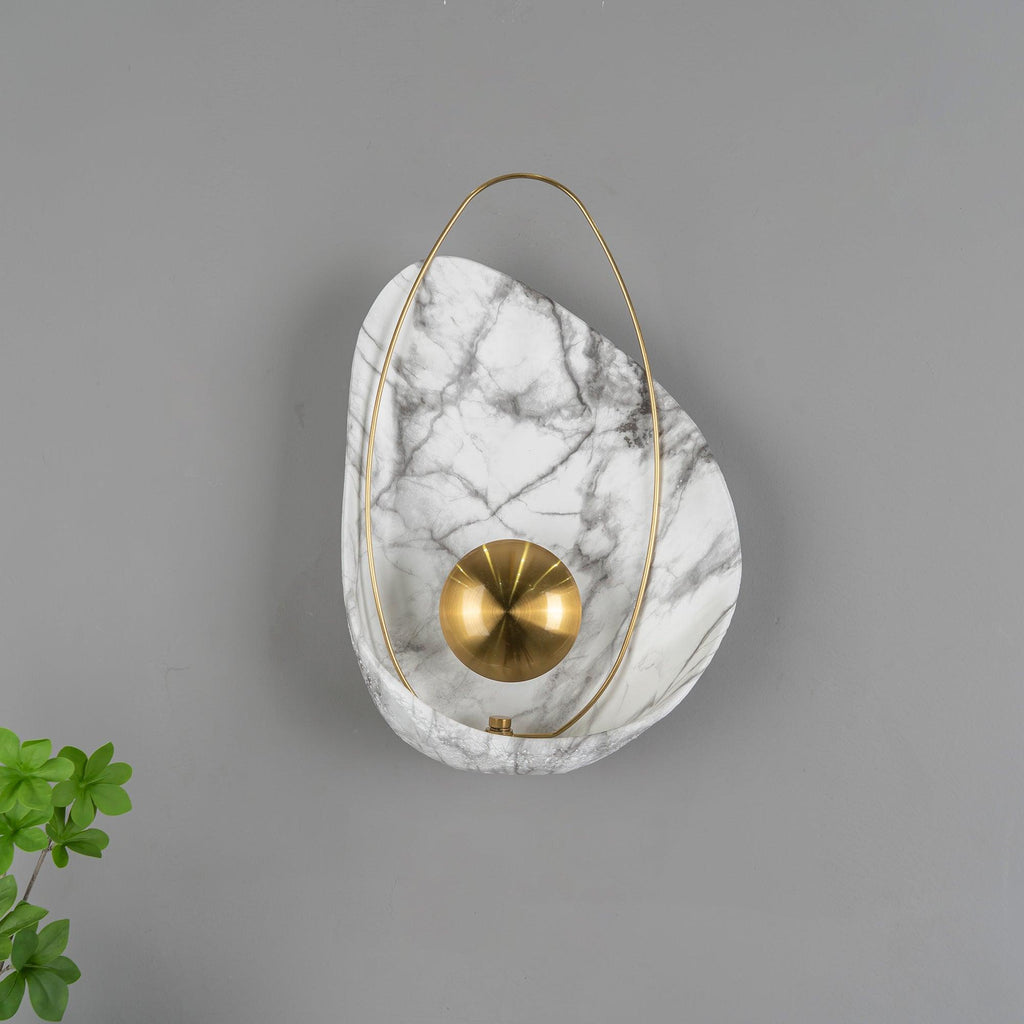 Unlit sconce on a grey wall, the brushed brass orb showing its radial finish, foliage at the left.