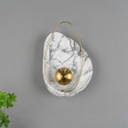 Unlit sconce on a grey wall, the brushed brass orb showing its radial finish, foliage at the left.