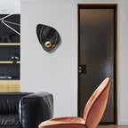 Black marbled sconce unlit on a white wall, beside a dark glass door and a velvet chair.