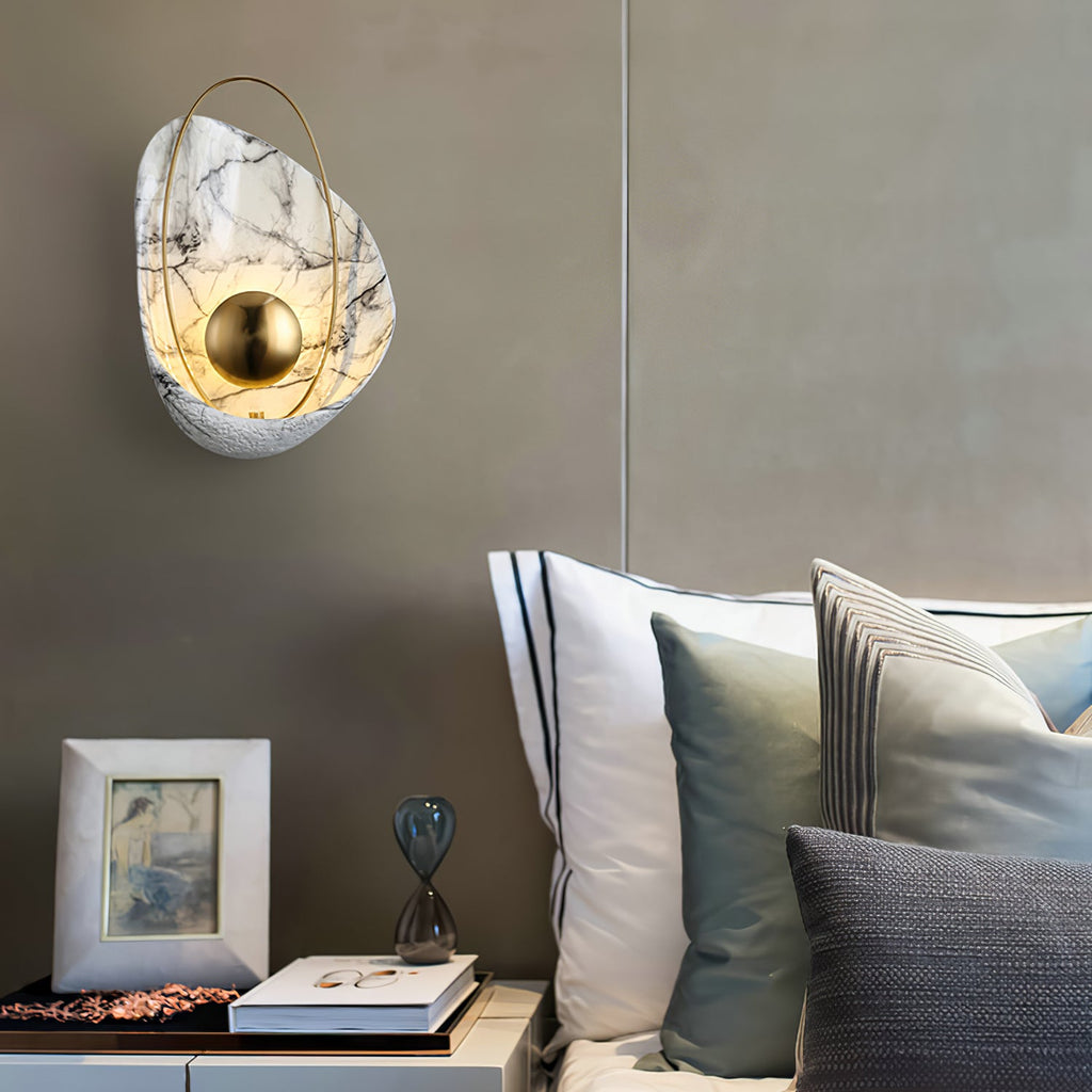 Lit sconce on a grey bedroom wall above a bedside table with a framed picture and an hourglass.