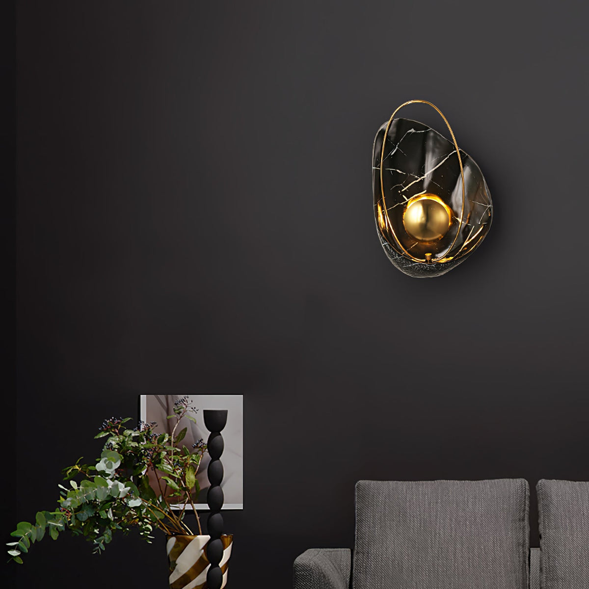 Black marbled sconce lit on a dark wall above a grey sofa, with a framed print and eucalyptus.