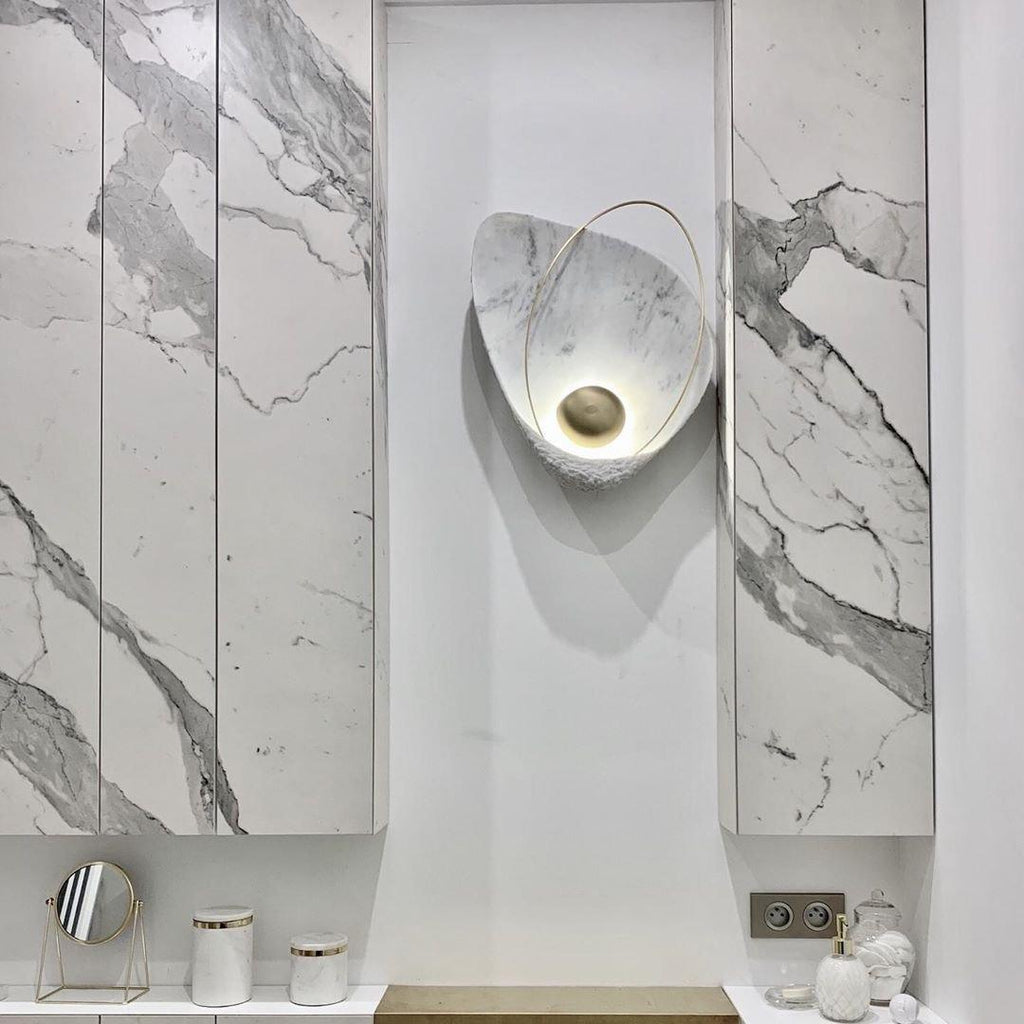 The lit sconce in a white recess between marble panels, above a vanity with a mirror and jars.