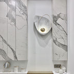 The lit sconce in a white recess between marble panels, above a vanity with a mirror and jars.