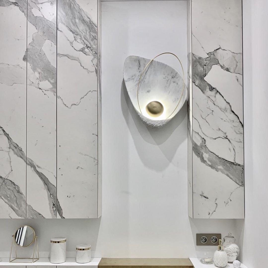 The lit sconce in a white recess between marble panels, above a vanity with a mirror and jars.