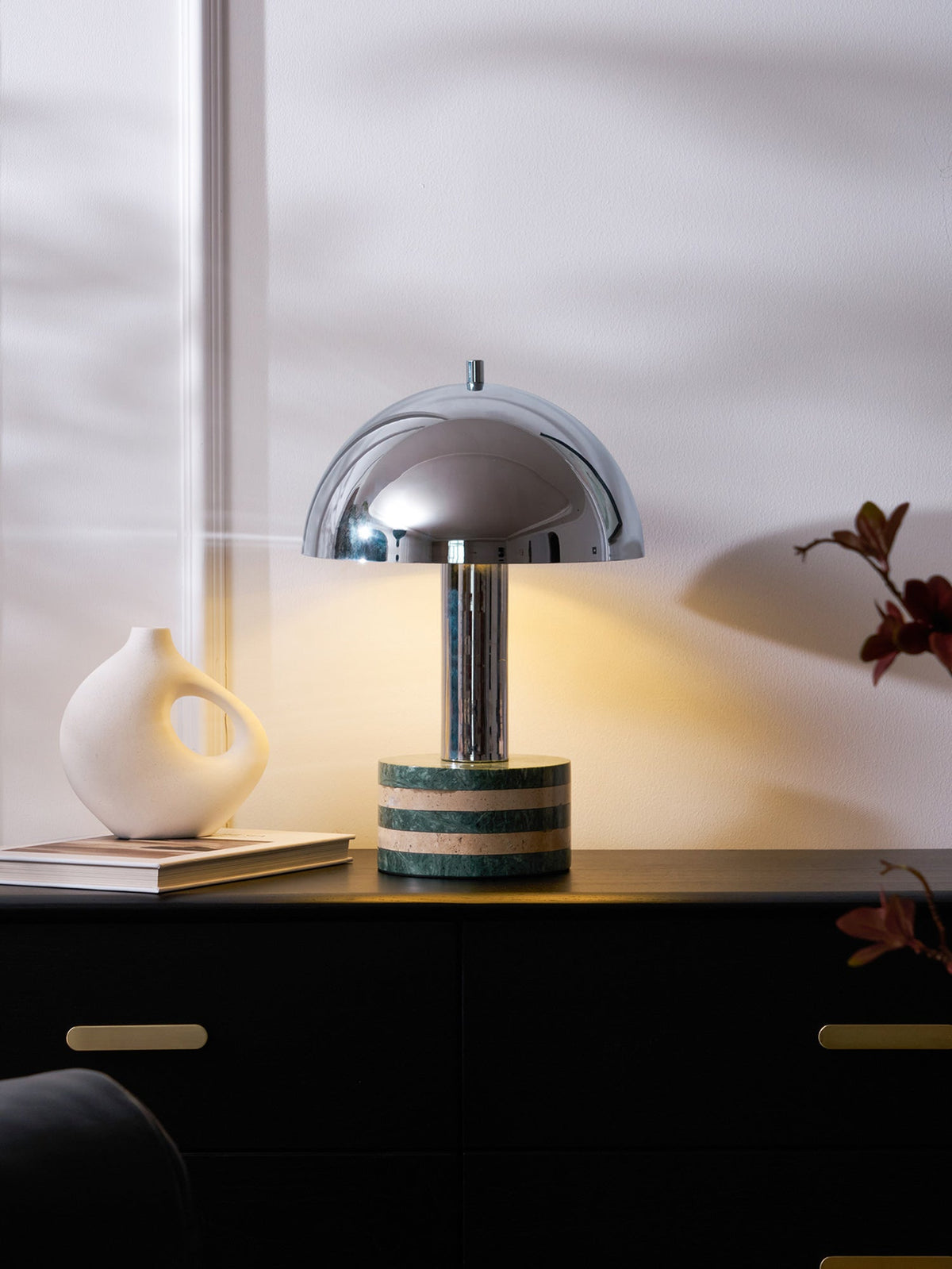 Close crop of the same Clérachic lamp glowing on the dark cabinet, brass drawer pulls and a paperback just visible below.