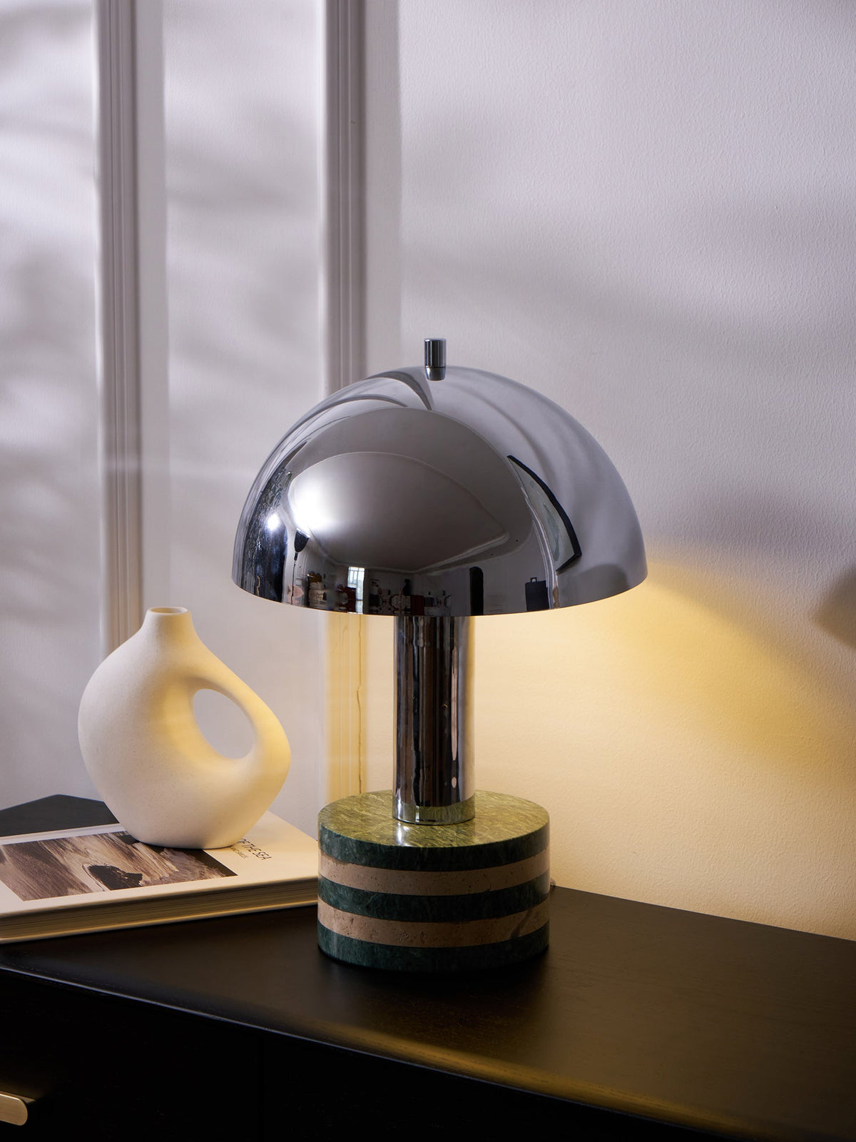Clérachic lamp lit beside the vase and book on the dresser, daylight coming through the window behind it.