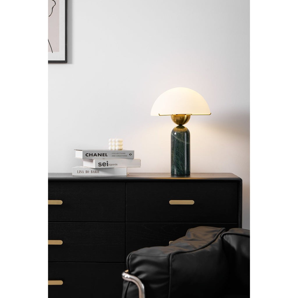Green marble Veviniel lamp lit on a black sideboard beside a leather chair and design books.