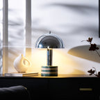 Clérachic table lamp lit beside a stack of books and a white ceramic vase on a dark cabinet, sheer curtain light behind it.