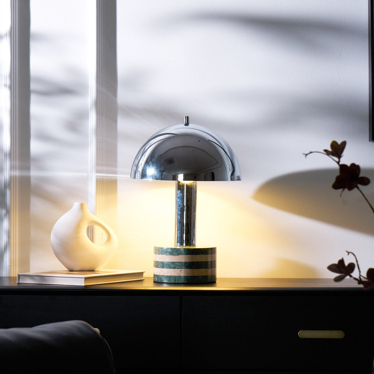 Clérachic table lamp lit beside a stack of books and a white ceramic vase on a dark cabinet, sheer curtain light behind it.