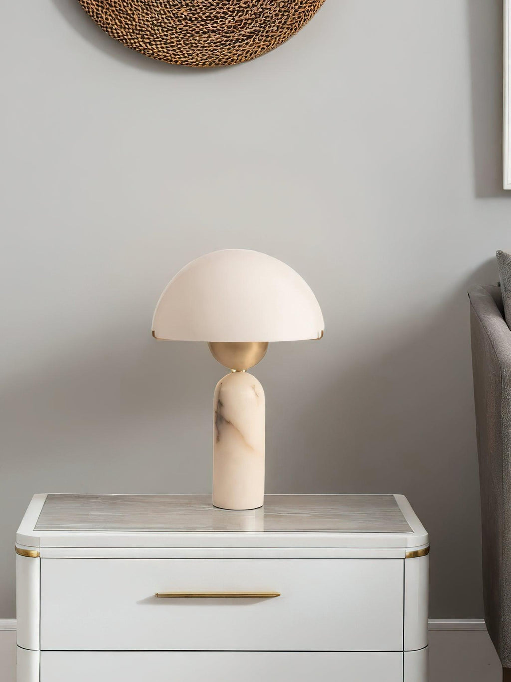 Veviniel lamp unlit on a white marble-topped nightstand, below a round woven wall hanging.