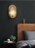 Lit on a black wall above a tan leather headboard, with a small plant and books alongside.
