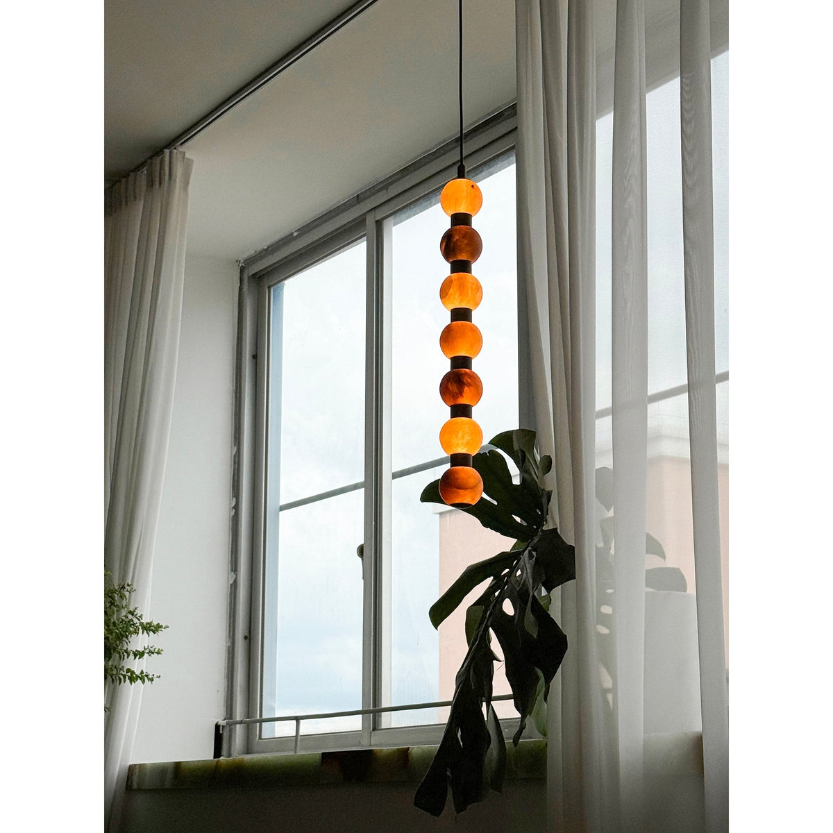 This Clérachic pendant glows deep orange against a bright window, its lower spheres near a monstera leaf.
