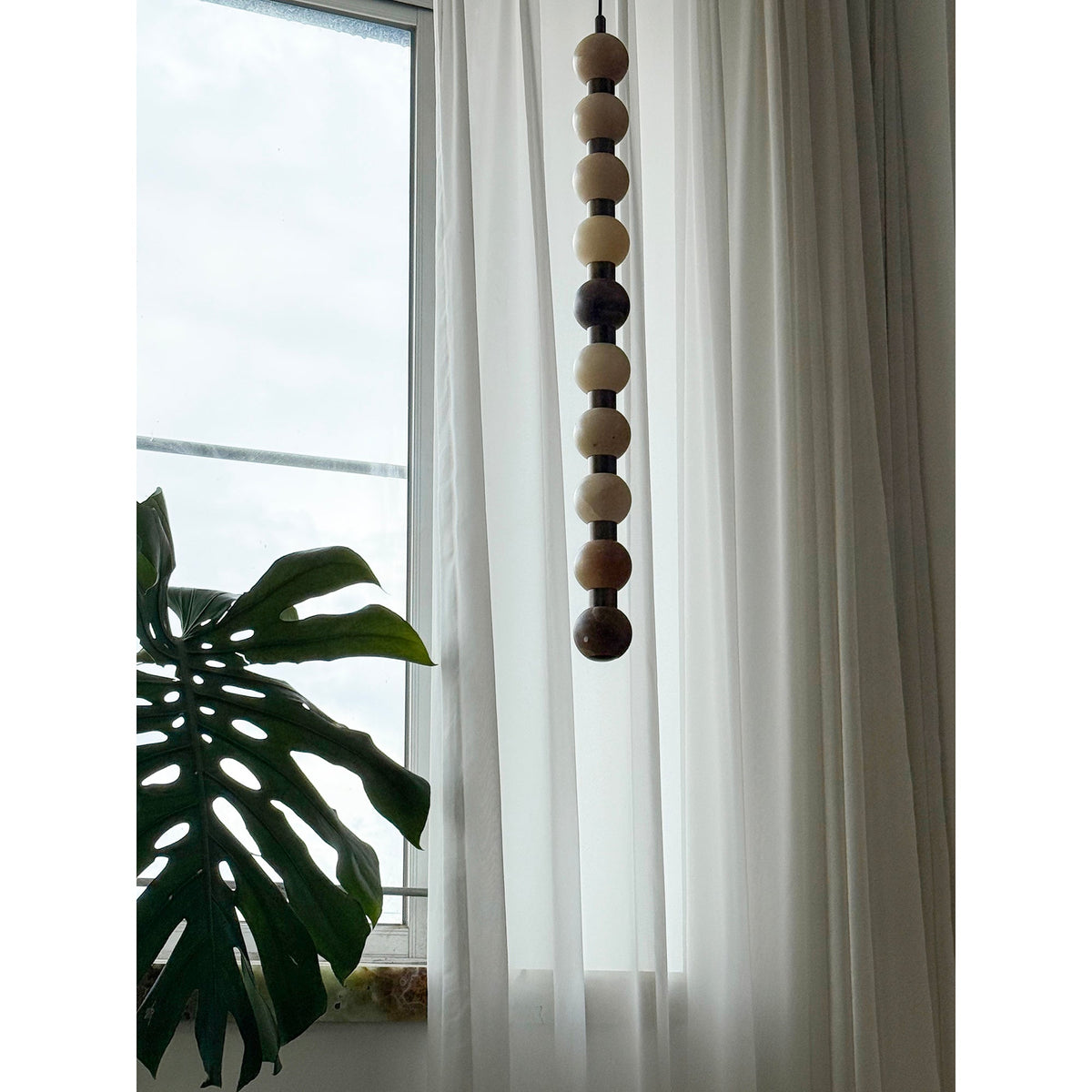 The pale natural stone tones of this Clérachic pendant hang beside a monstera leaf and sheer curtain.