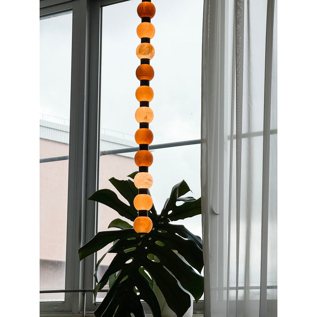 This Clérachic alabaster sphere pendant hangs against a window, a broad monstera leaf reaching up below it.