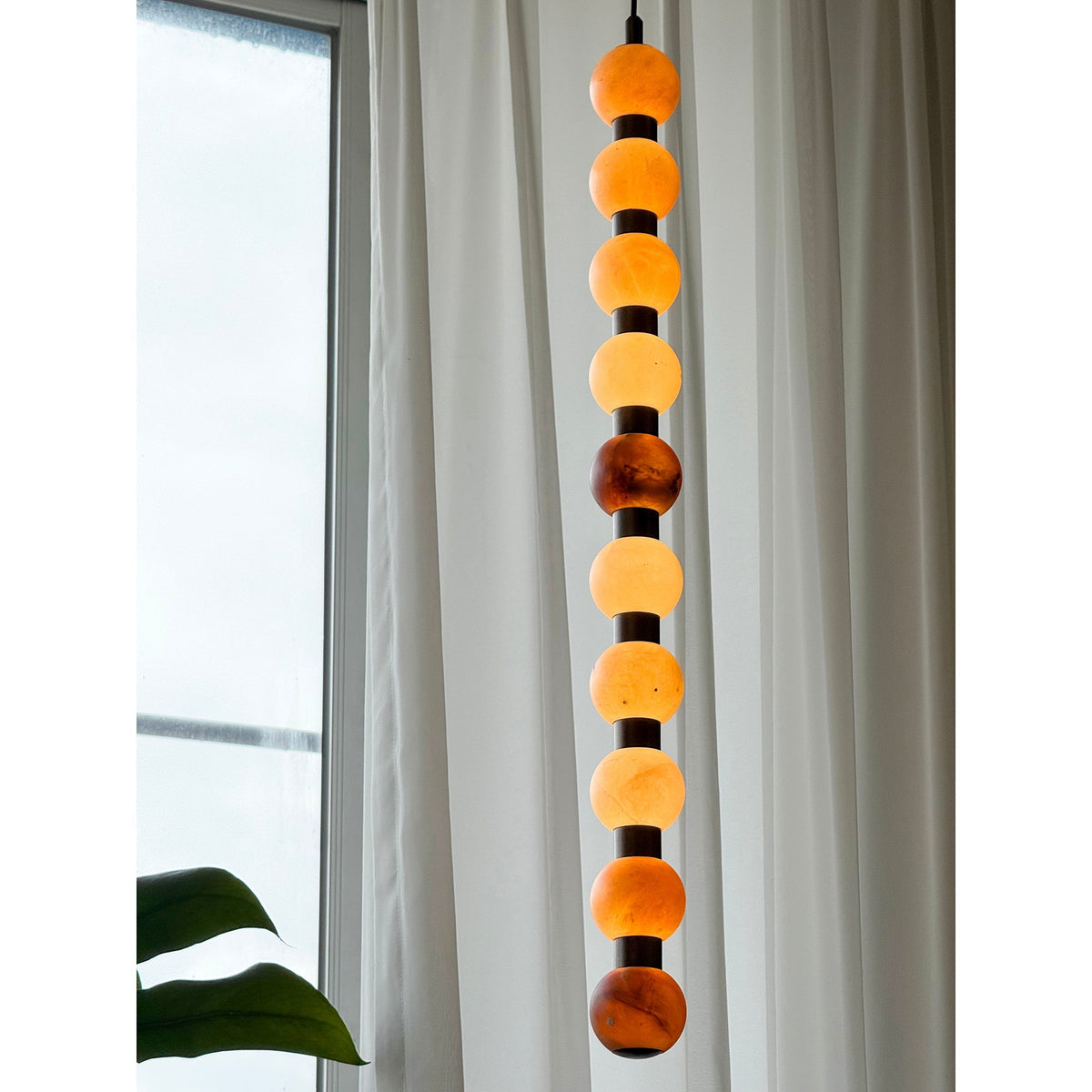 The full length of this Clérachic pendant glows warm against a window, ending near a monstera leaf.