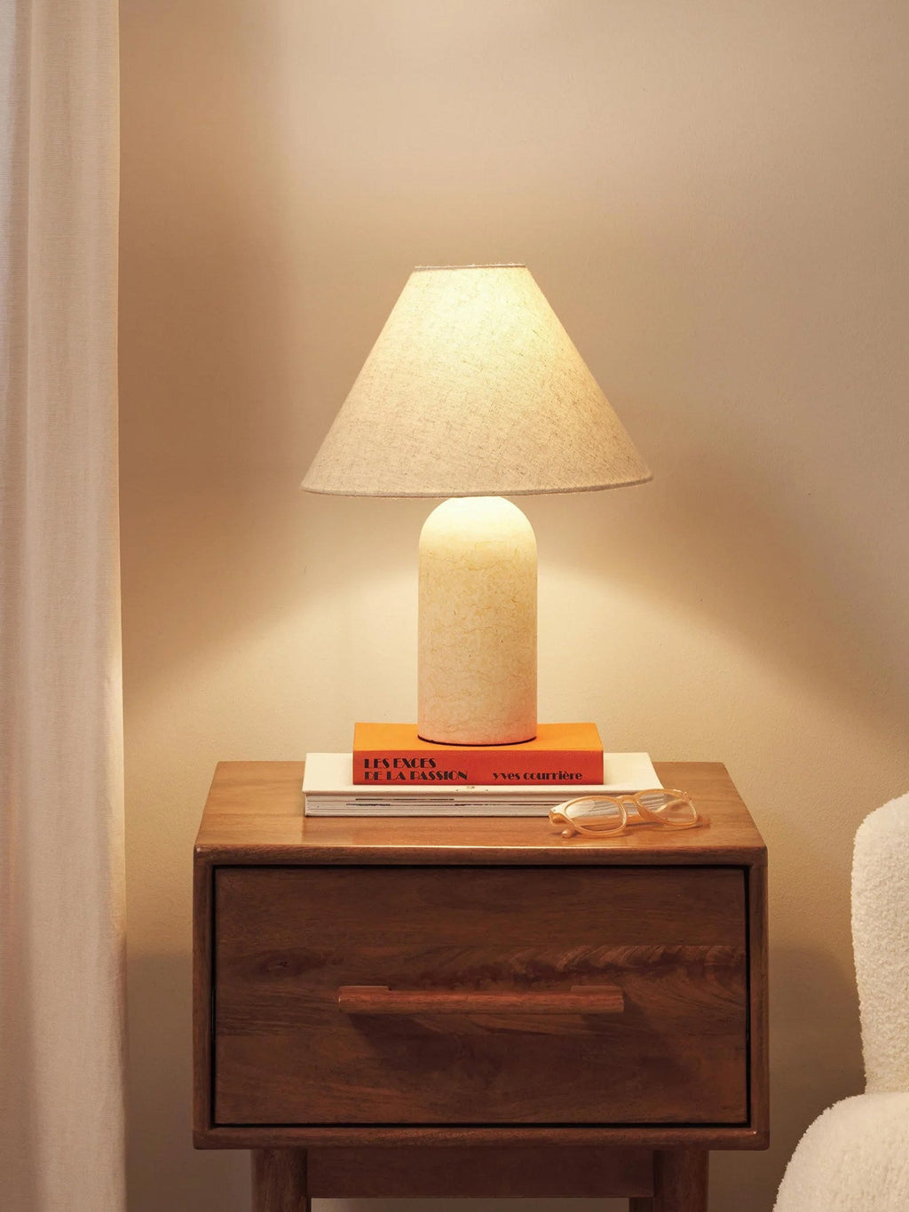 Clérachic lamp in the pale cream marble finish, glowing on a nightstand stacked with books beside eyeglasses.