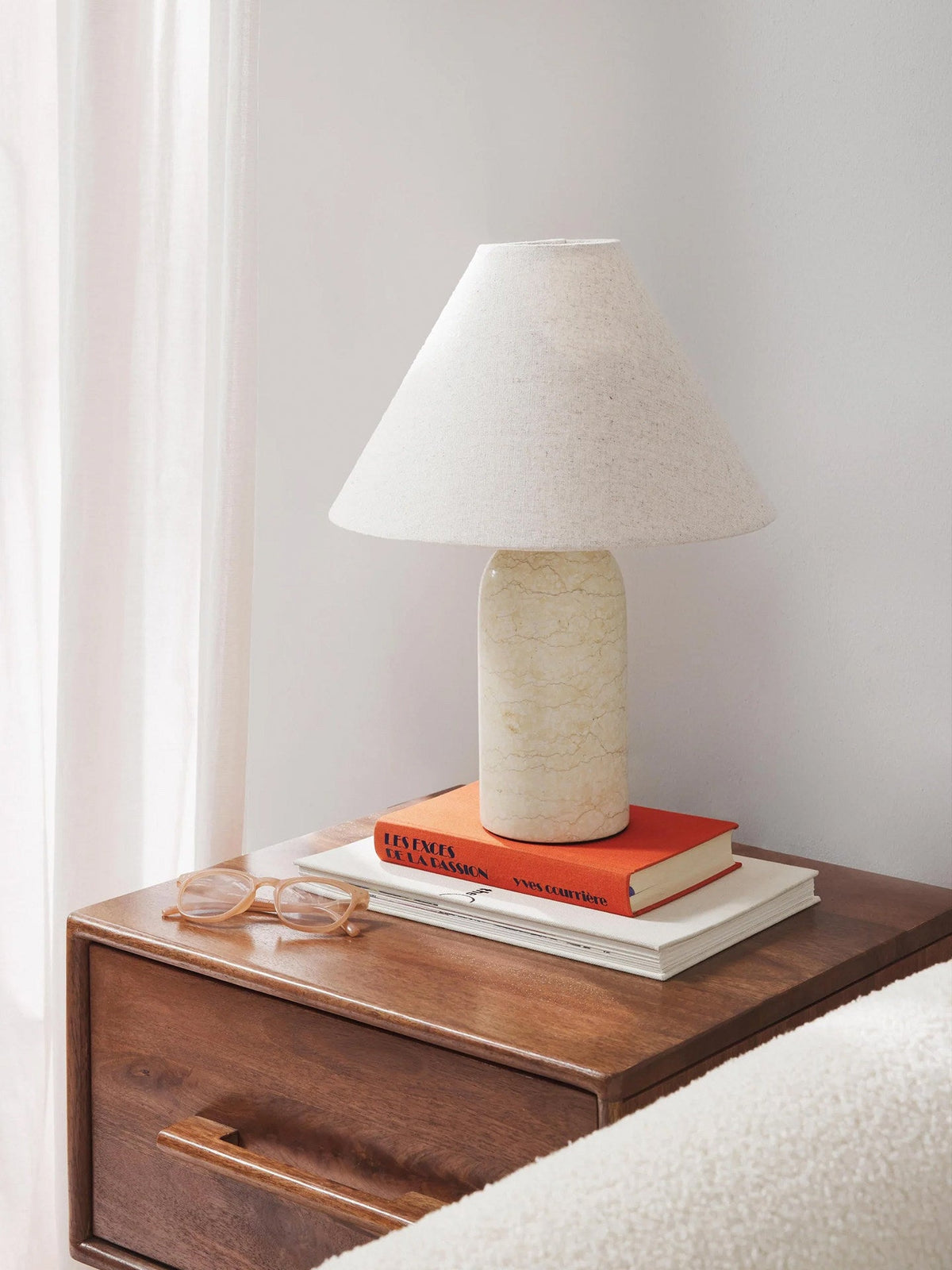 Clérachic lamp in a pale cream marble finish, lit on a walnut nightstand beside eyeglasses and stacked books.