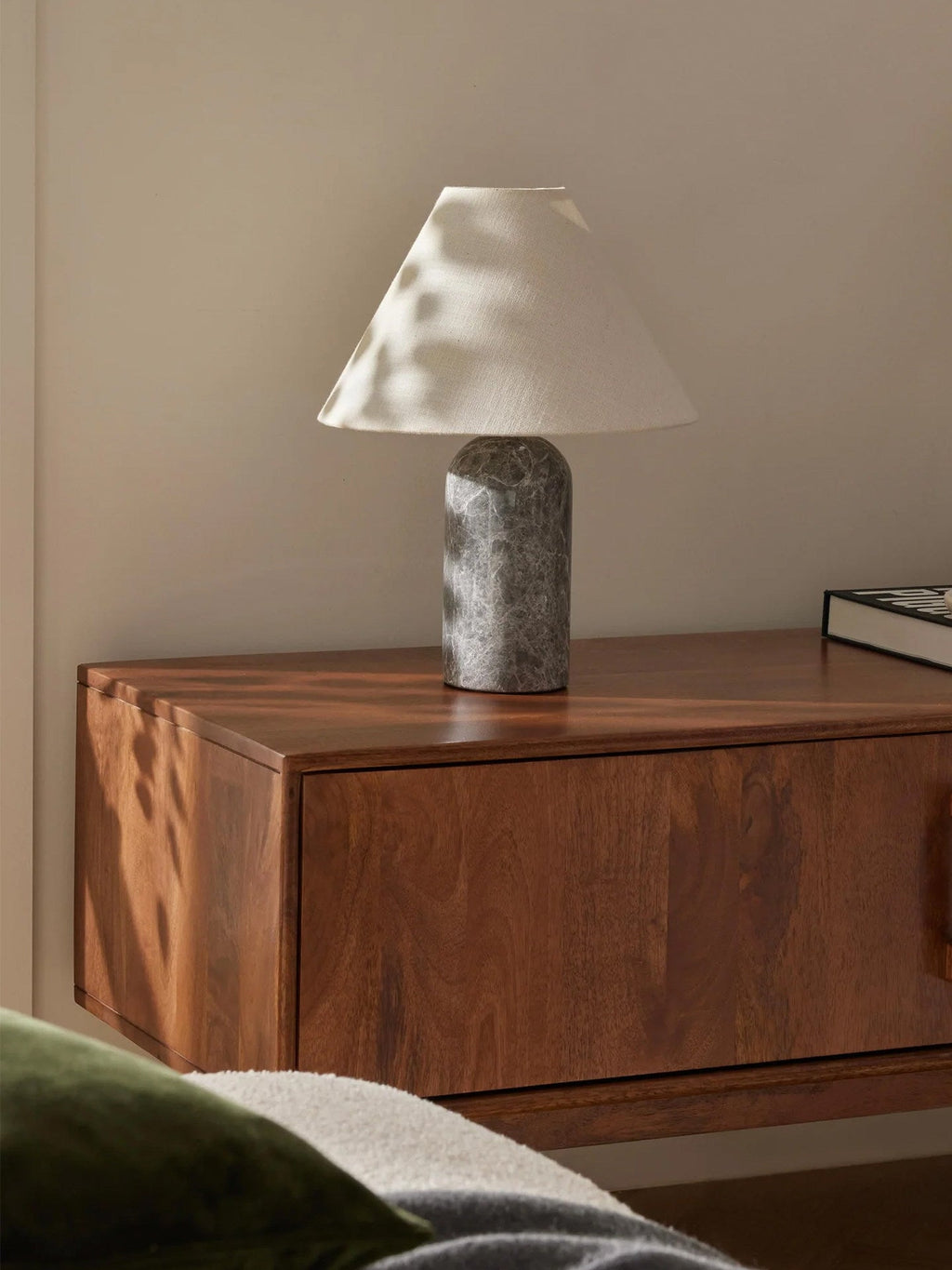 Clérachic lamp in a grey marble finish, lit on a floating wooden nightstand beside a green cushion.