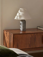 Clérachic lamp in a grey marble finish, lit on a floating wooden nightstand beside a green cushion.