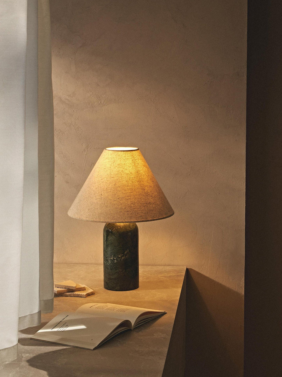 Clérachic lamp in a dark green marble finish, glowing in a dim alcove beside an open magazine on the floor.