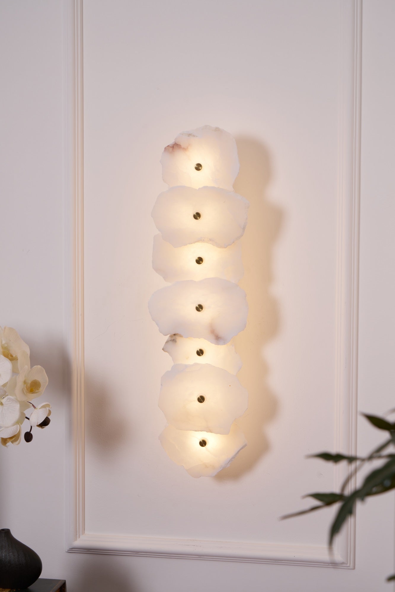 Six-head Alaeila alabaster wall light lit on a white panelled wall, orchid stem in the corner.