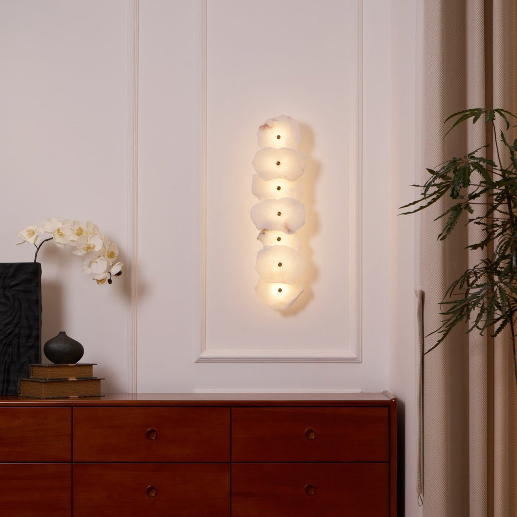 Wide room view of the lit alabaster wall lamp above a wooden sideboard beside a curtained window.