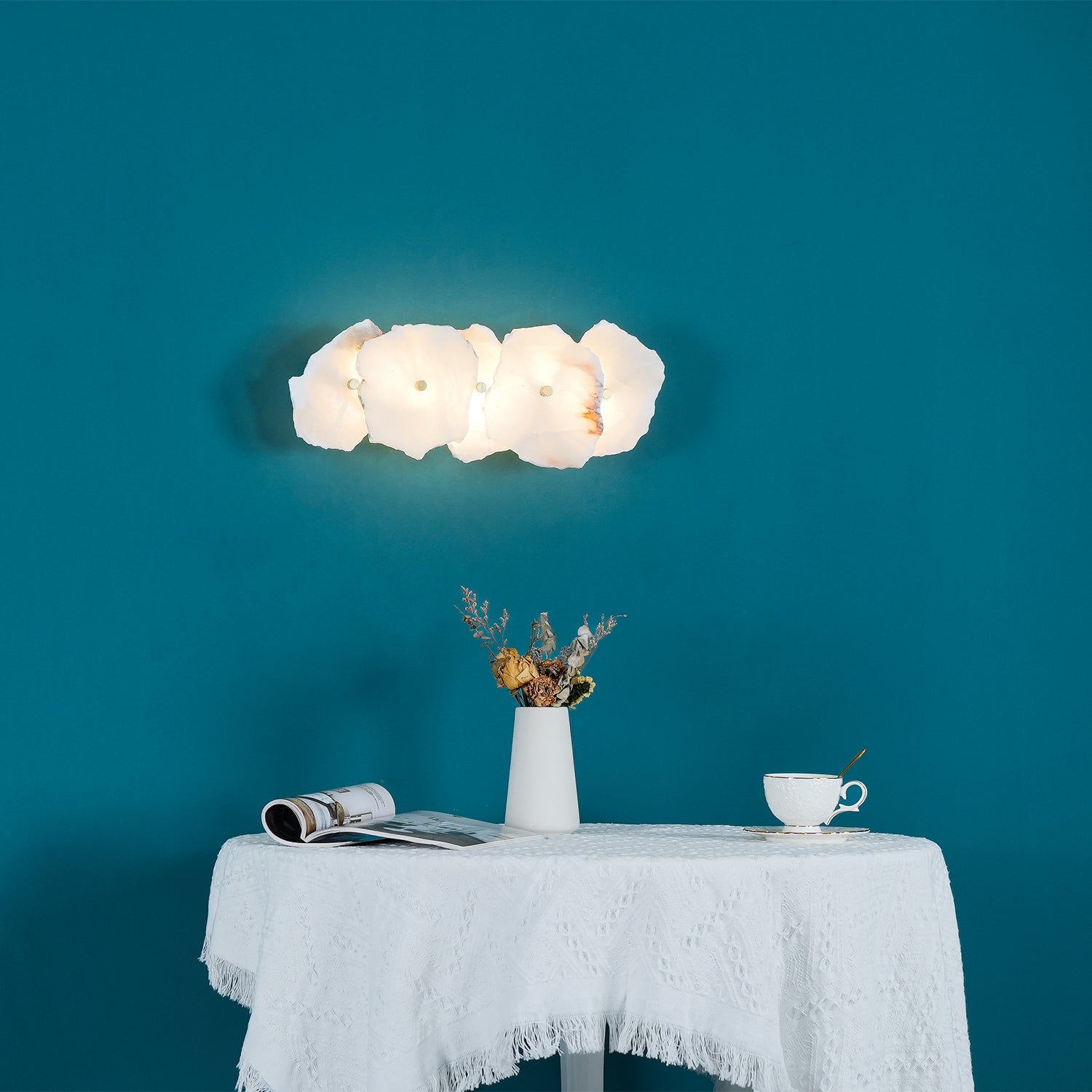 Horizontal five-head alabaster wall lamp lit above a lace-covered table on a deep teal wall.