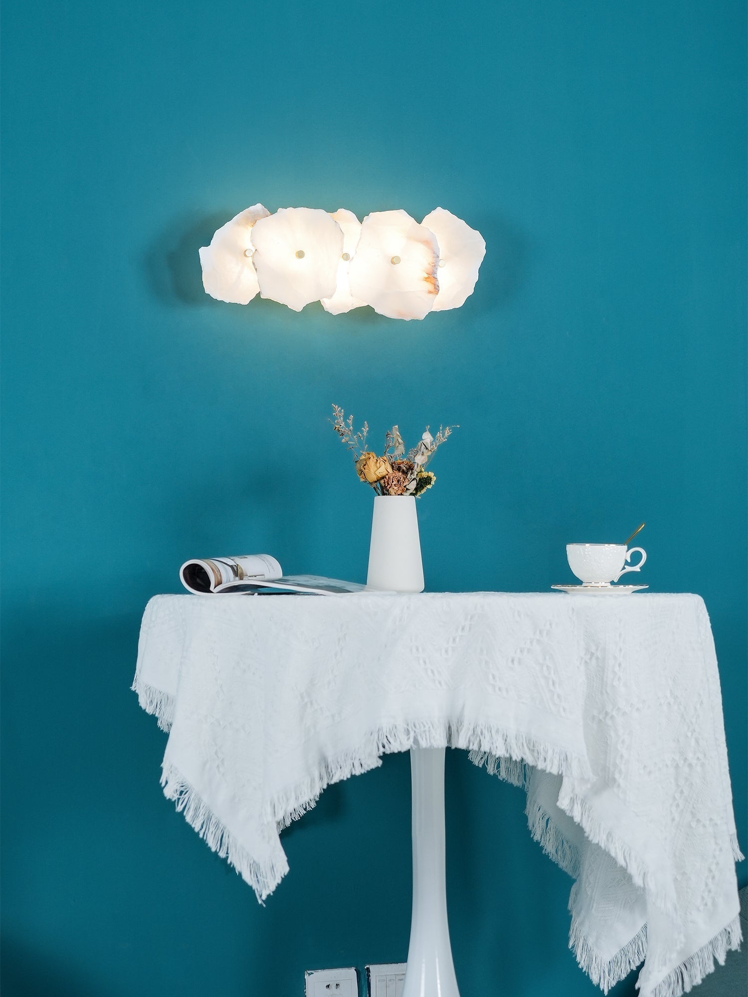 Teal-walled corner with the wide Alaeila alabaster wall light lit above a dried-flower vase.
