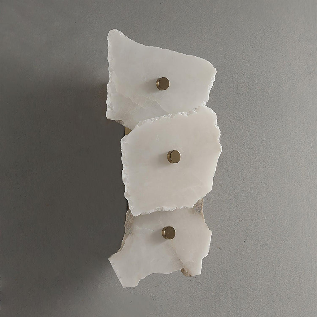 Unlit three-head alabaster wall lamp on a grey wall, chipped stone edges and brass studs in daylight.