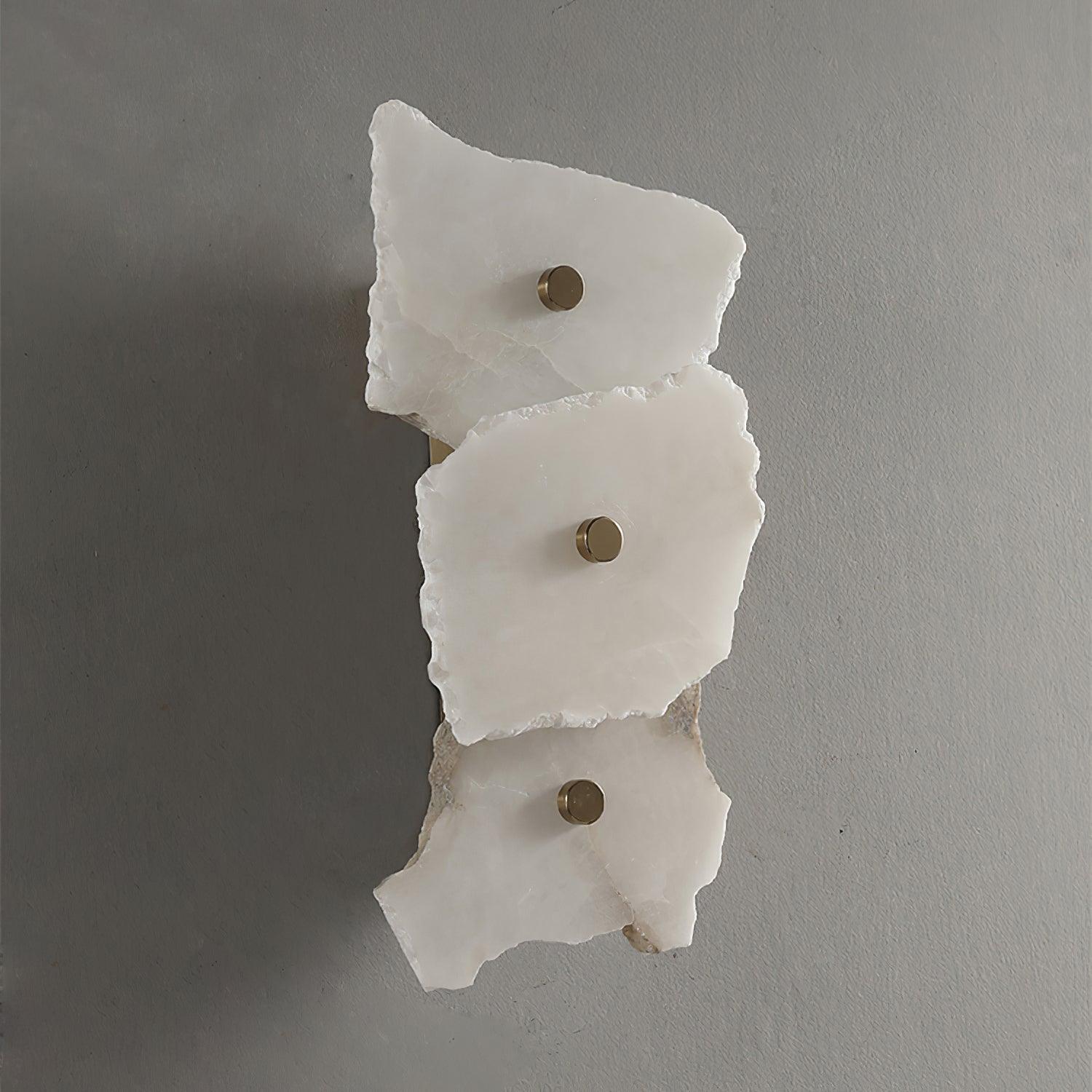 Unlit three-head alabaster wall lamp on a grey wall, chipped stone edges and brass studs in daylight.