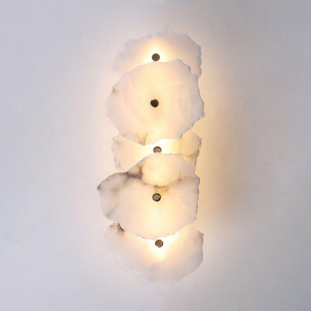 Five-head alabaster wall lamp close up on a pale wall, warm light pooling behind each stone slab.
