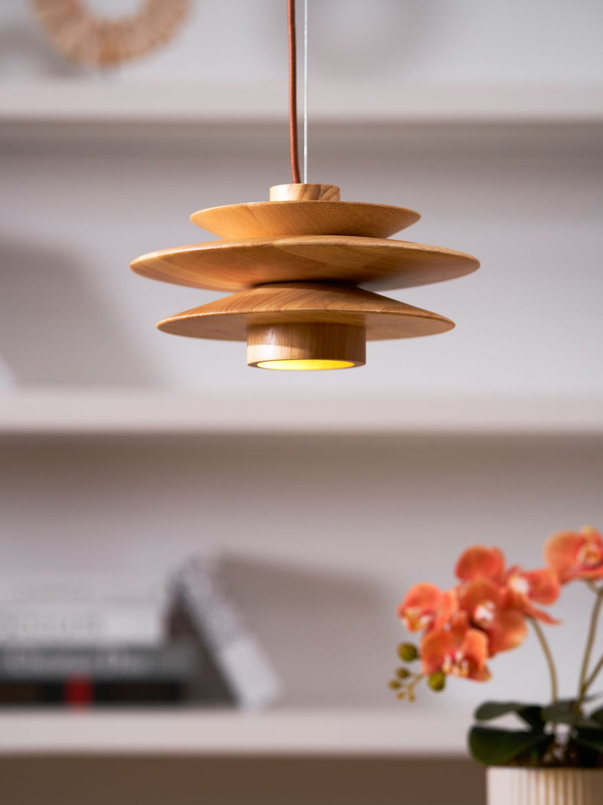 Glowing warmly above a desk, this Clérachic pendant in nature wood has an orchid and bookshelf softly blurred behind it.