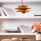 This Clérachic pendant in nature wood hangs above a white bookshelf holding books and an orange orchid, with an open book on the desk below.