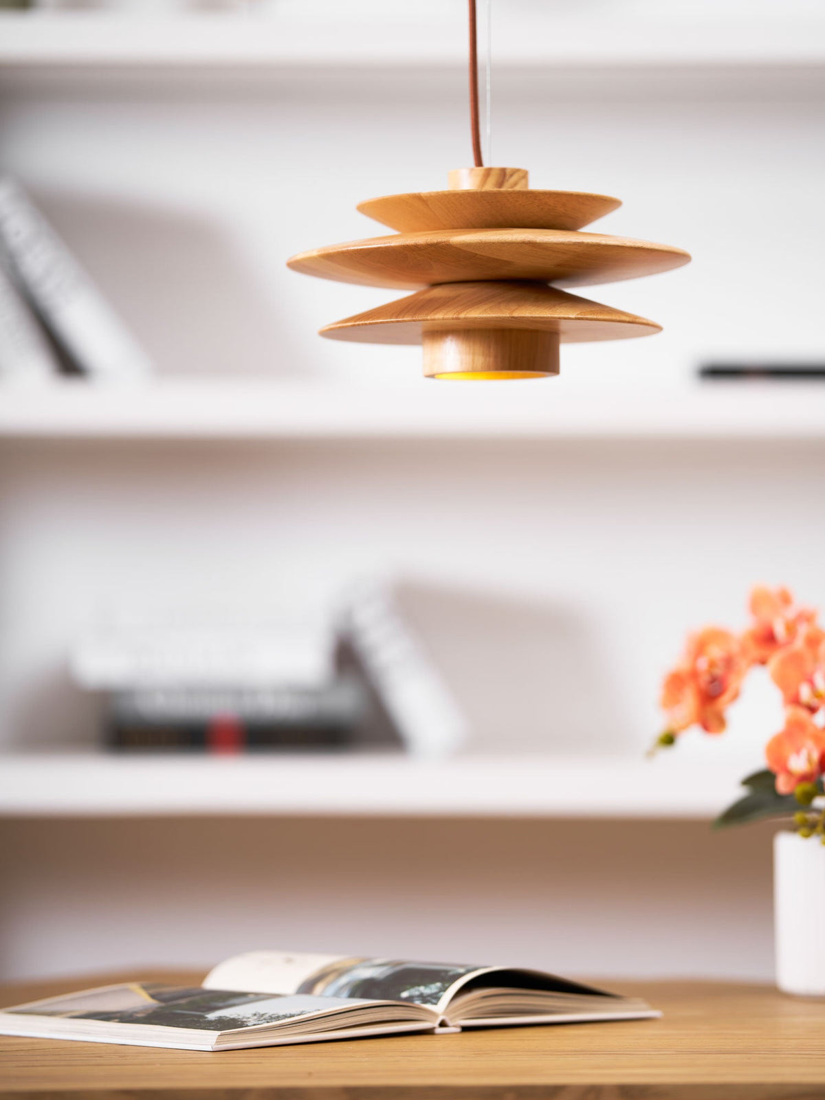 Seen closer, this Clérachic pendant in nature wood hangs near a white bookshelf stacked with books and a magazine.